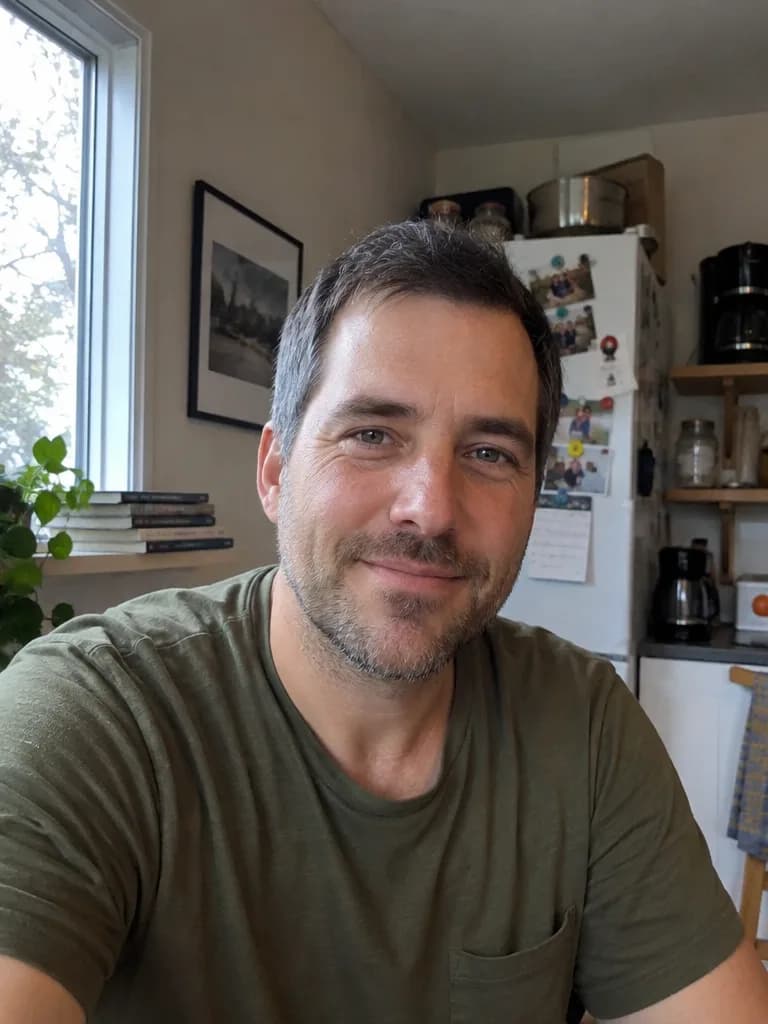 Foto casual de um homem com cabelos grisalhos nas têmporas e barba por fazer, usando camiseta verde-oliva à mesa da cozinha
