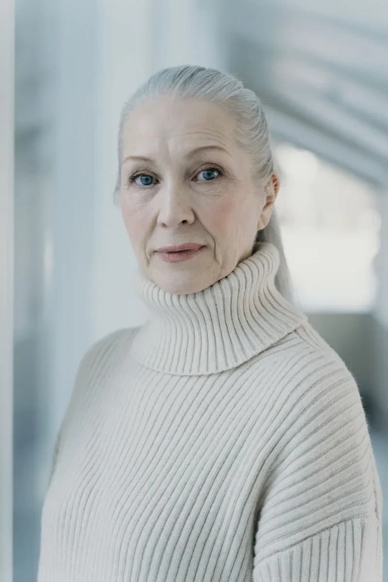 Older woman with grey hair in a white turtleneck by a window