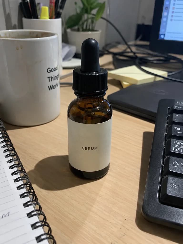 杂乱办公桌上的琥珀色精华瓶,旁边有键盘、线和杯子,顶光阴影明显